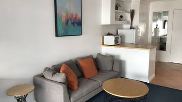 A modern living room with a grey sofa, two round coffee tables, a kitchen area with white cabinets, and a colorful abstract painting on the wall.