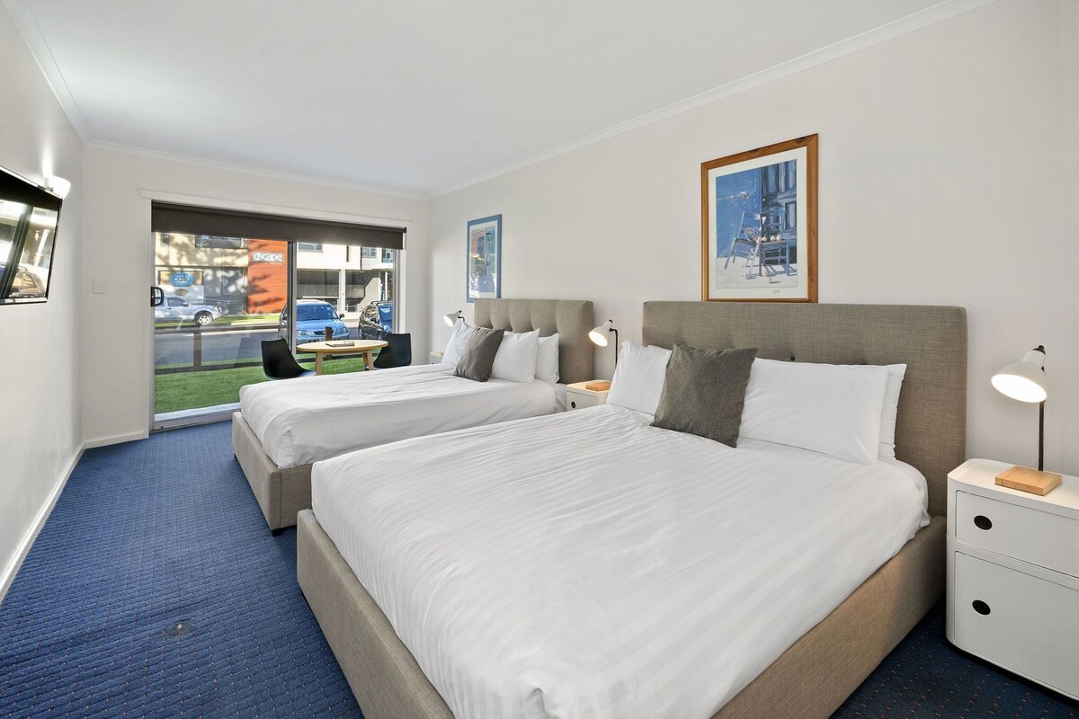 A hotel room with two beds, a small table, a chair, and a window with a view of the street.