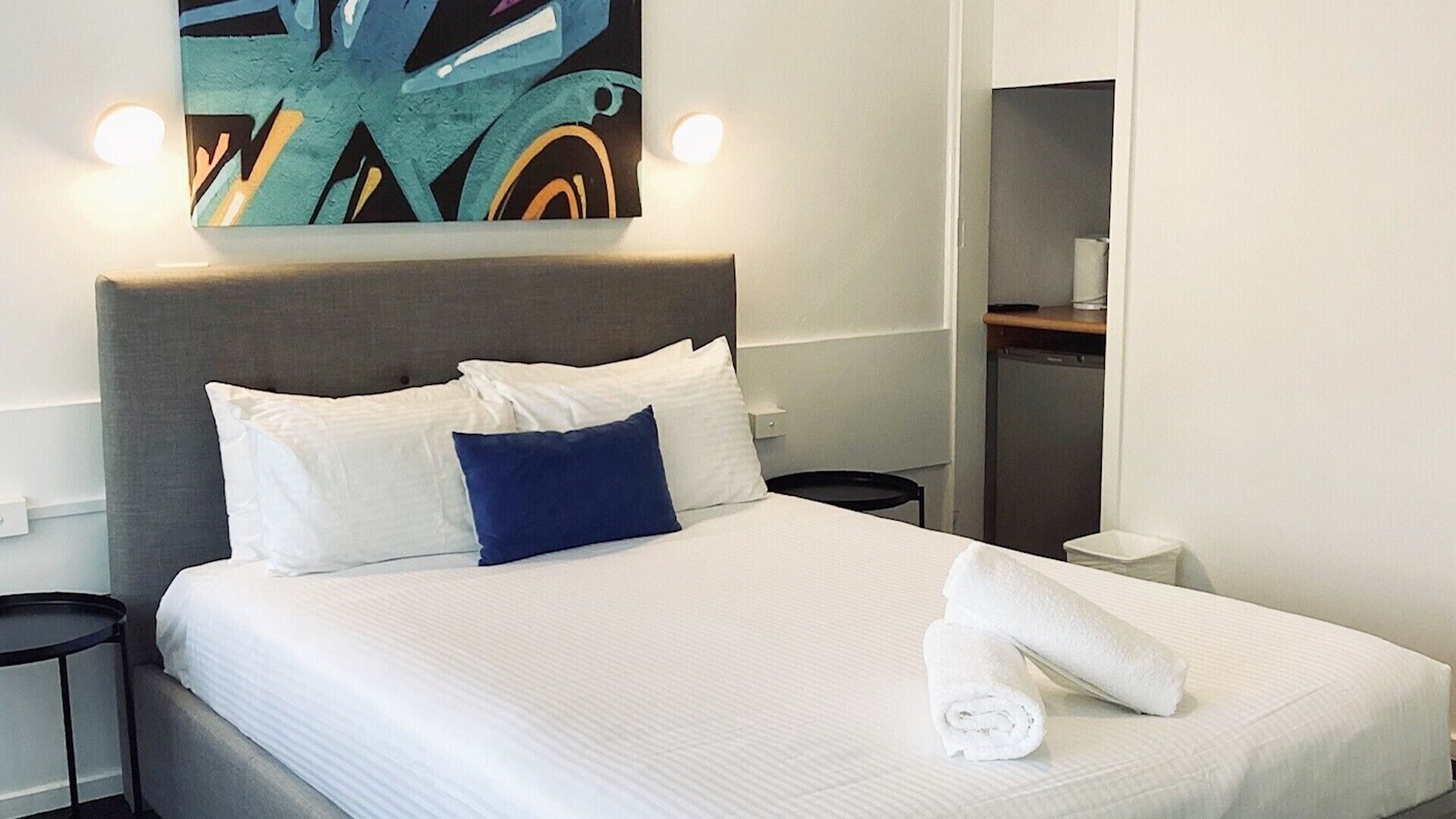 A hotel room with a bed, a nightstand, a towel, and a painting on the wall.