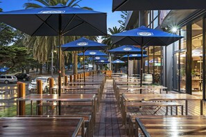 Mesas de madera al aire libre y sombrillas azules con un logotipo, dispuestas en una terraza con vista a palmeras y una calle.