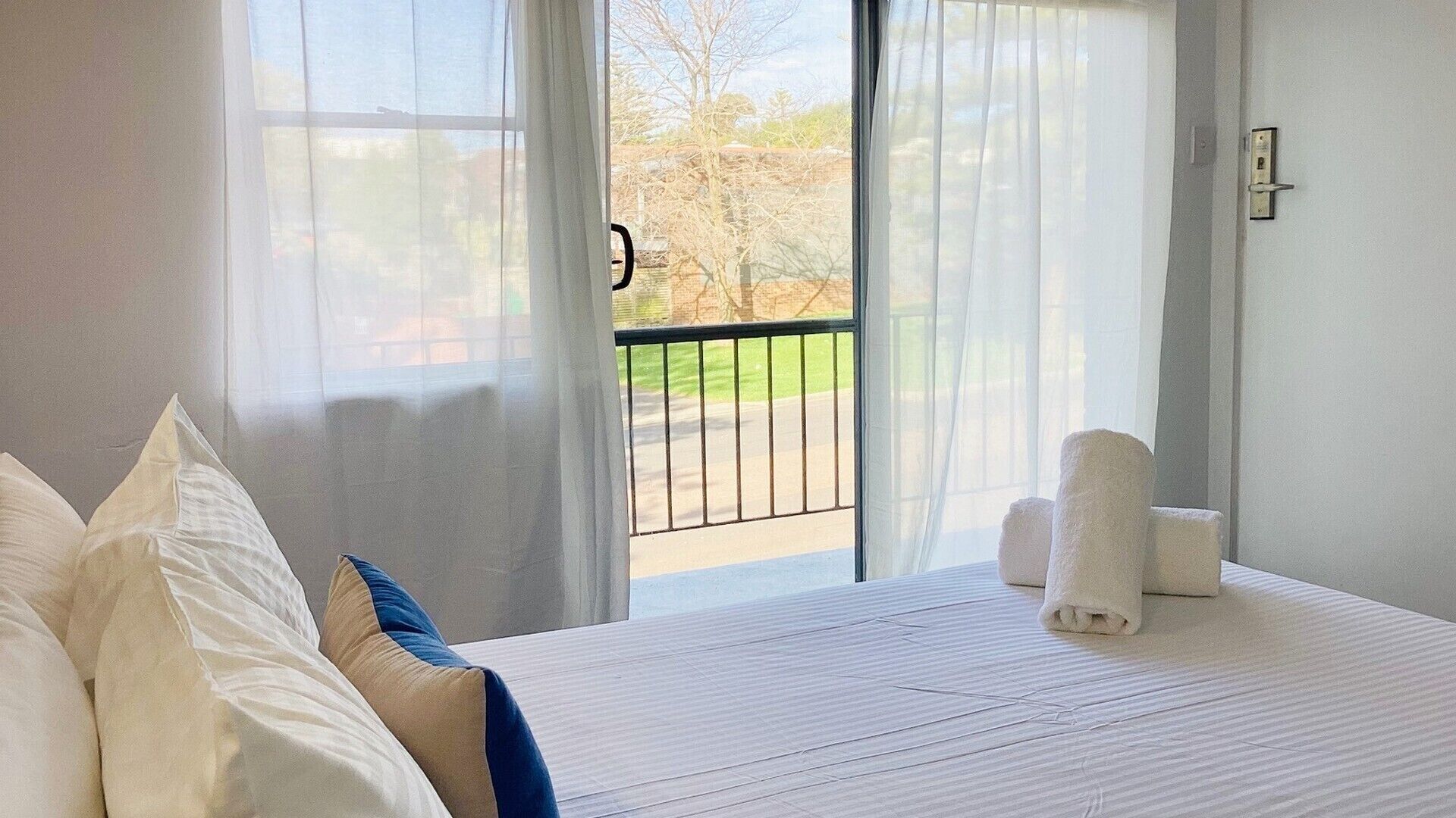 A hotel room with a bed, a view of a balcony, and a door.