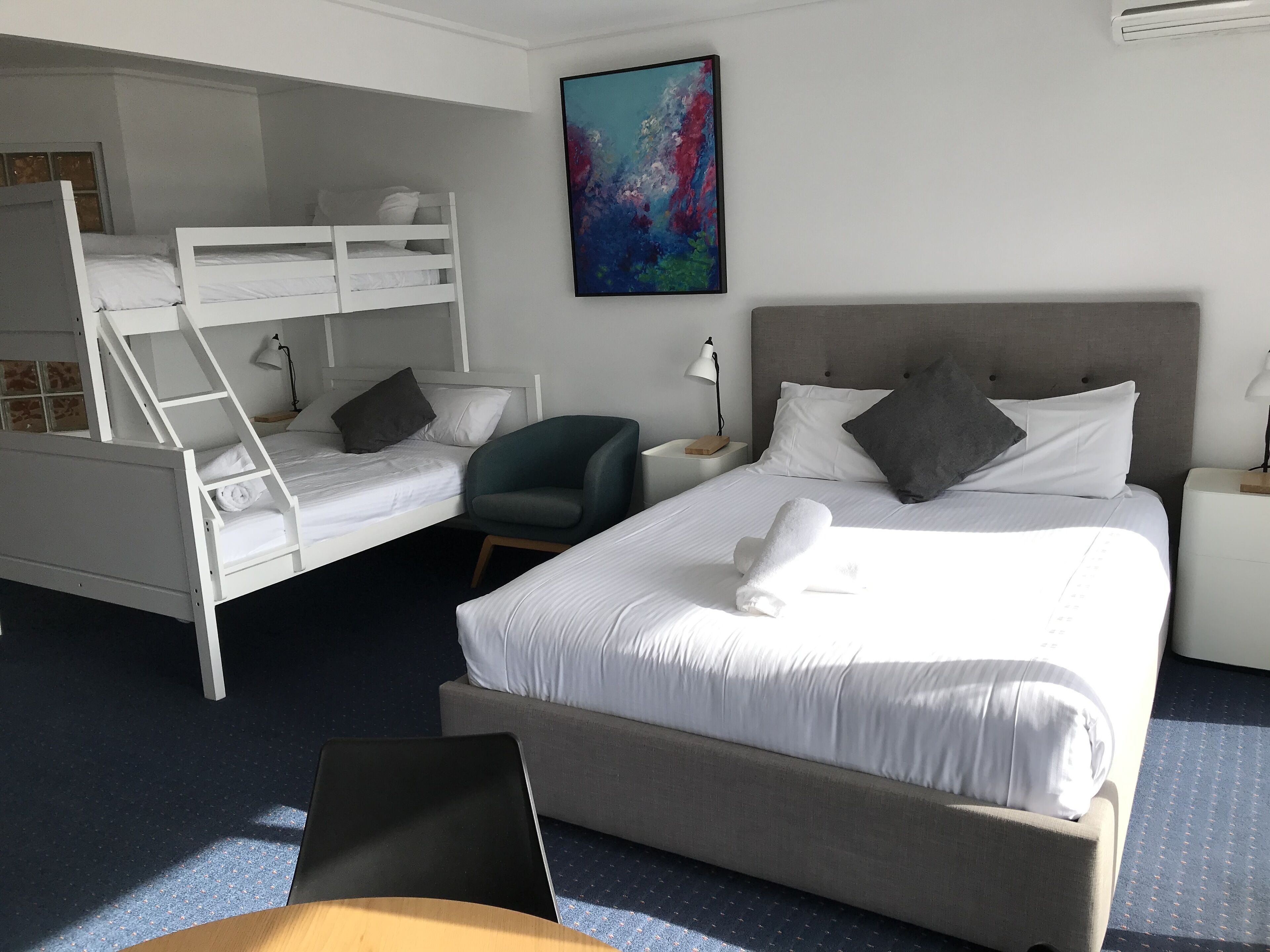 Ein Hotelzimmer mit Etagenbett, einem Einzelbett, einem Stuhl, einem Schreibtisch und einem Bild an der Wand.