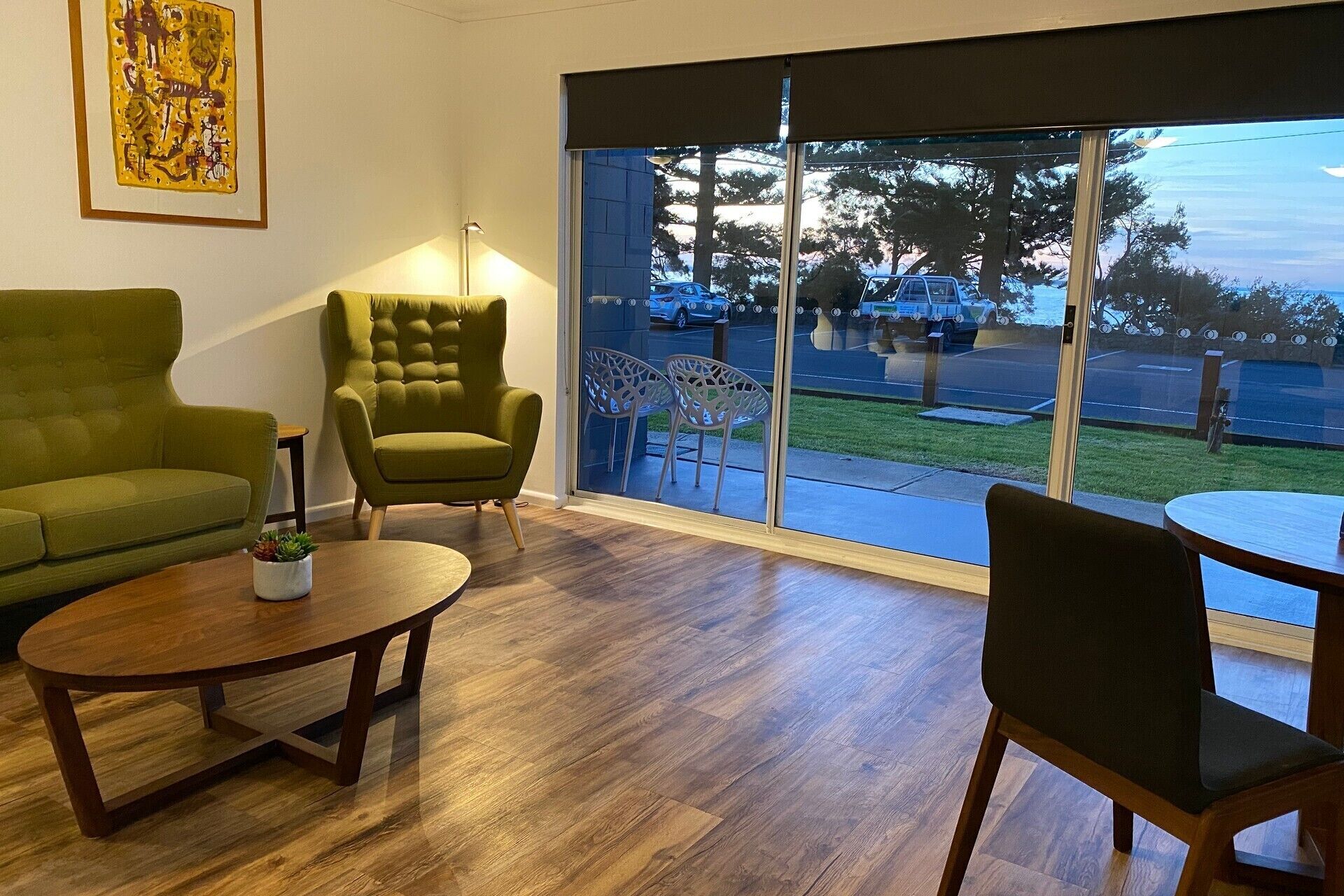 A room with a green sofa, a tufted armchair, and a round wooden table. There is a large window with a view of a parking lot and trees.