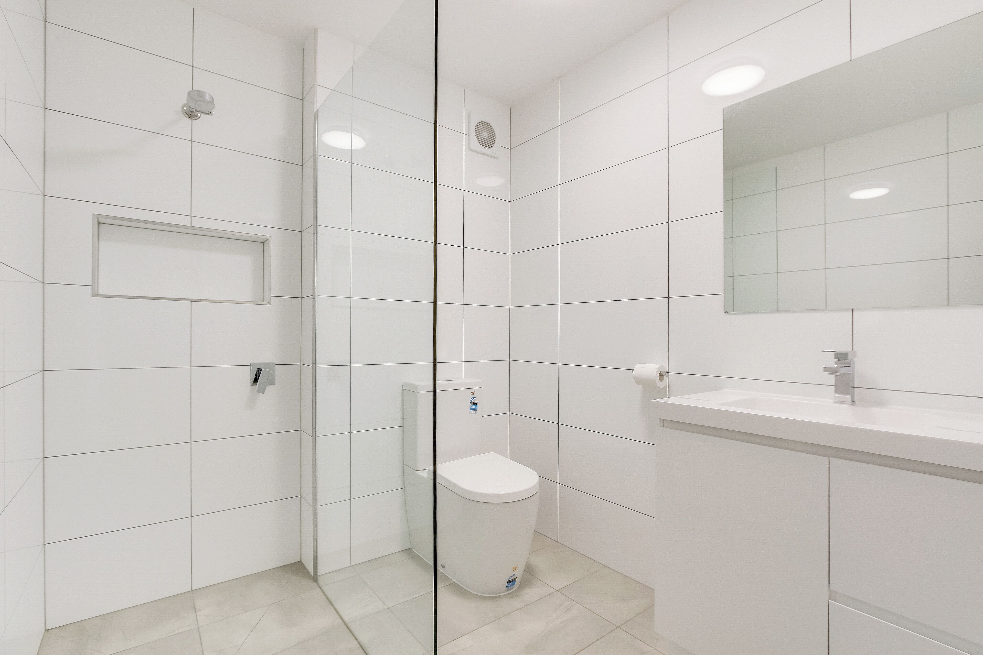 A modern bathroom with white tiles, a glass shower enclosure, a toilet, and a sink with a mirror.