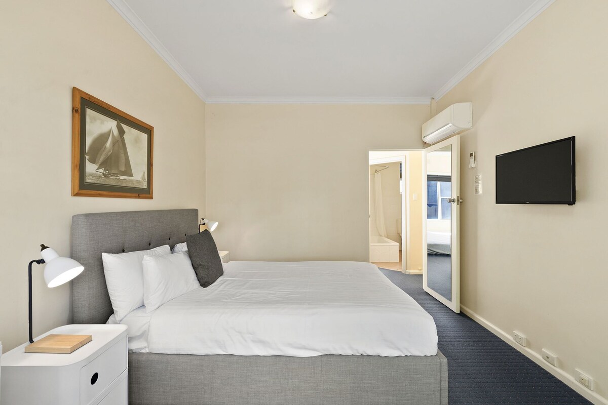Una habitación de hotel con cama, lámpara de noche, mesita de noche, un cuadro enmarcado y televisión.