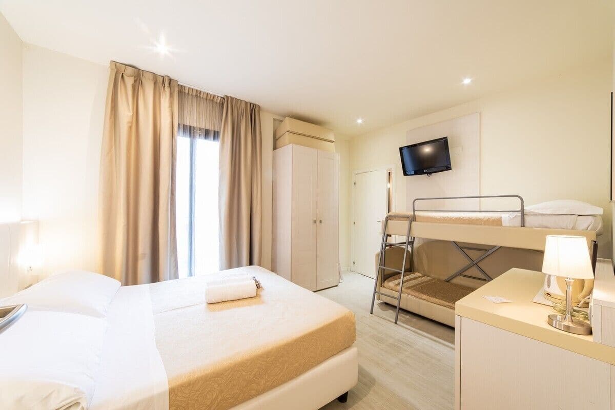 Habitación de hotel con cama individual, literas, escritorio con lámpara, televisor en la pared y armario.