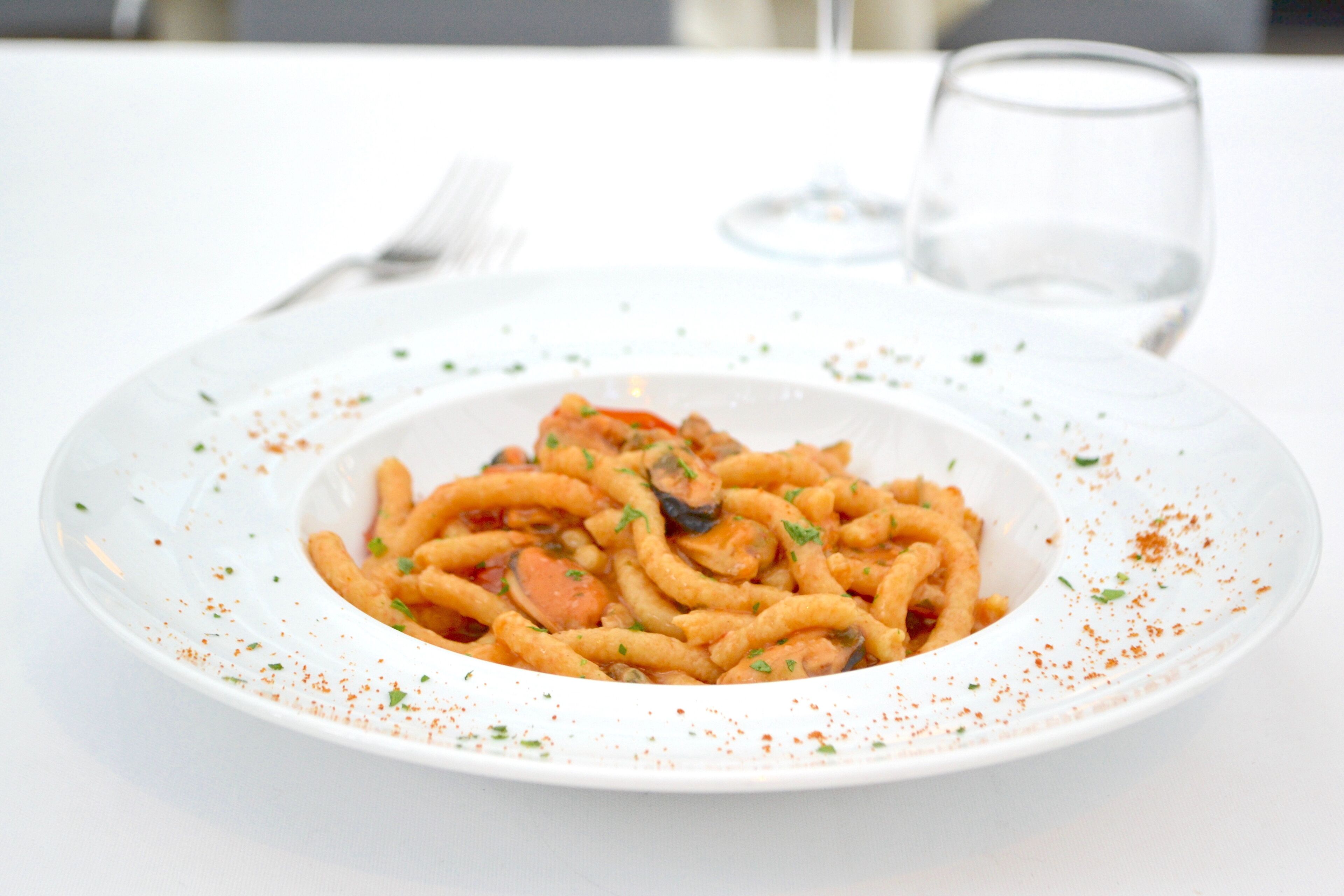 A plate of pasta with a garnish, a fork, and a glass of water on a white table.