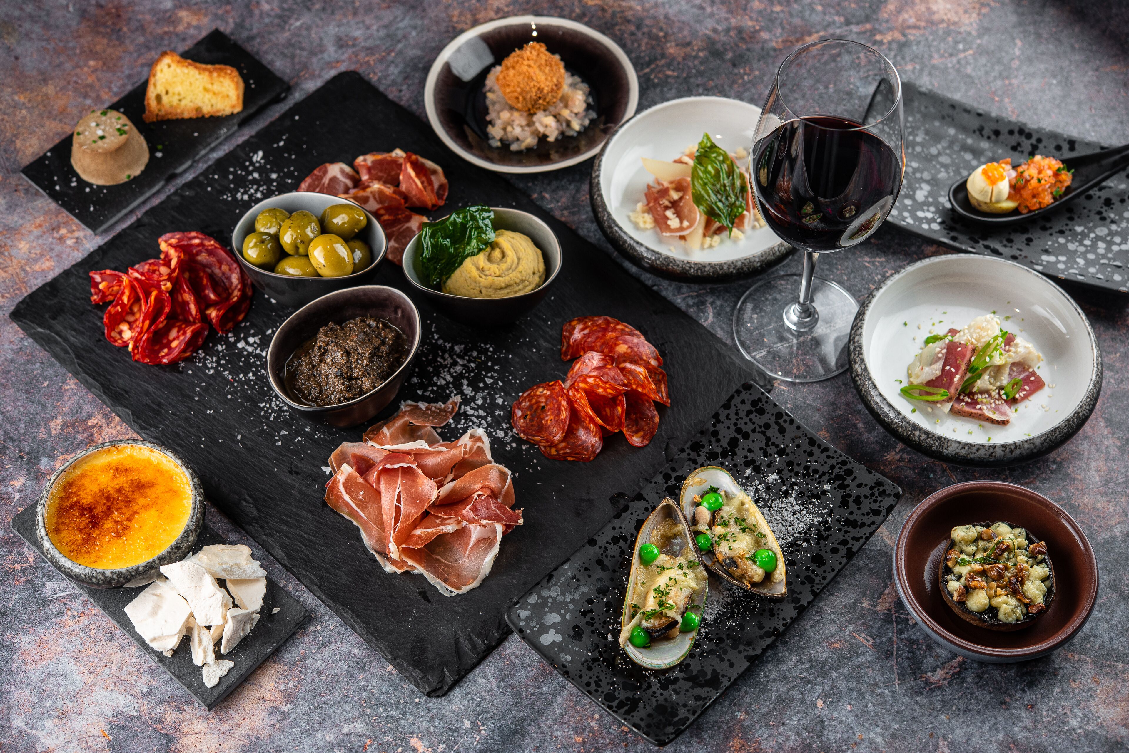 A variety of food items and a glass of red wine on a dark slate table.