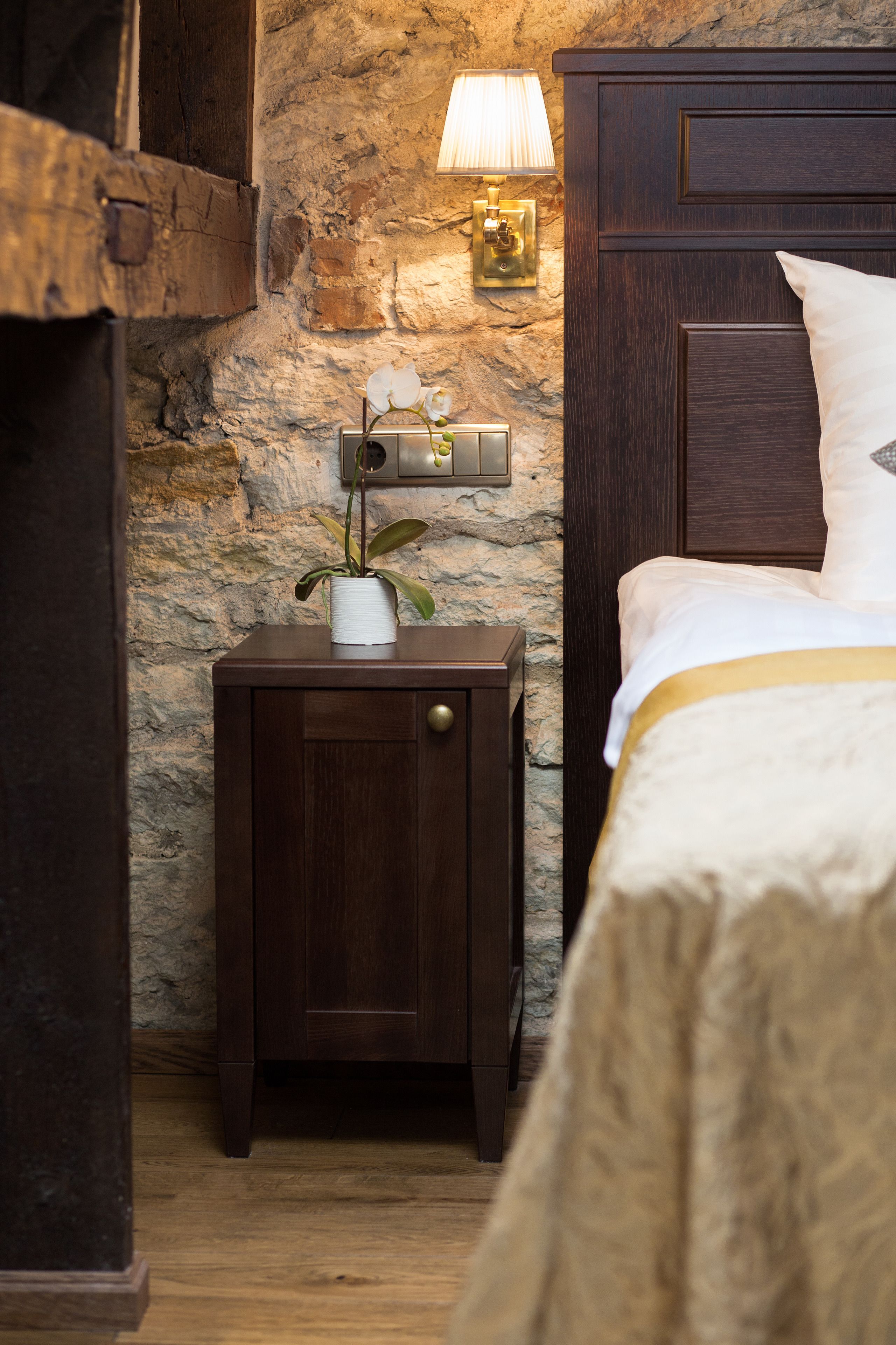 Un dormitorio con pared de piedra, una mesita de noche con lámpara y una cama con almohada blanca.