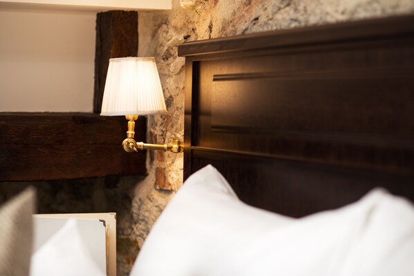 A close-up of a wall-mounted lamp with a white shade, a wooden headboard, and a stone wall.