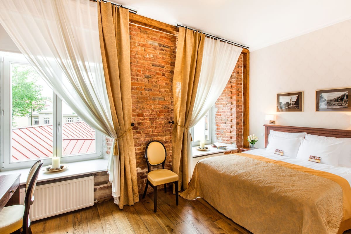 Une chambre d’hôtel avec un mur en briques, un plancher en bois, un lit avec une literie blanche, une chaise, un bureau et une fenêtre avec des rideaux.