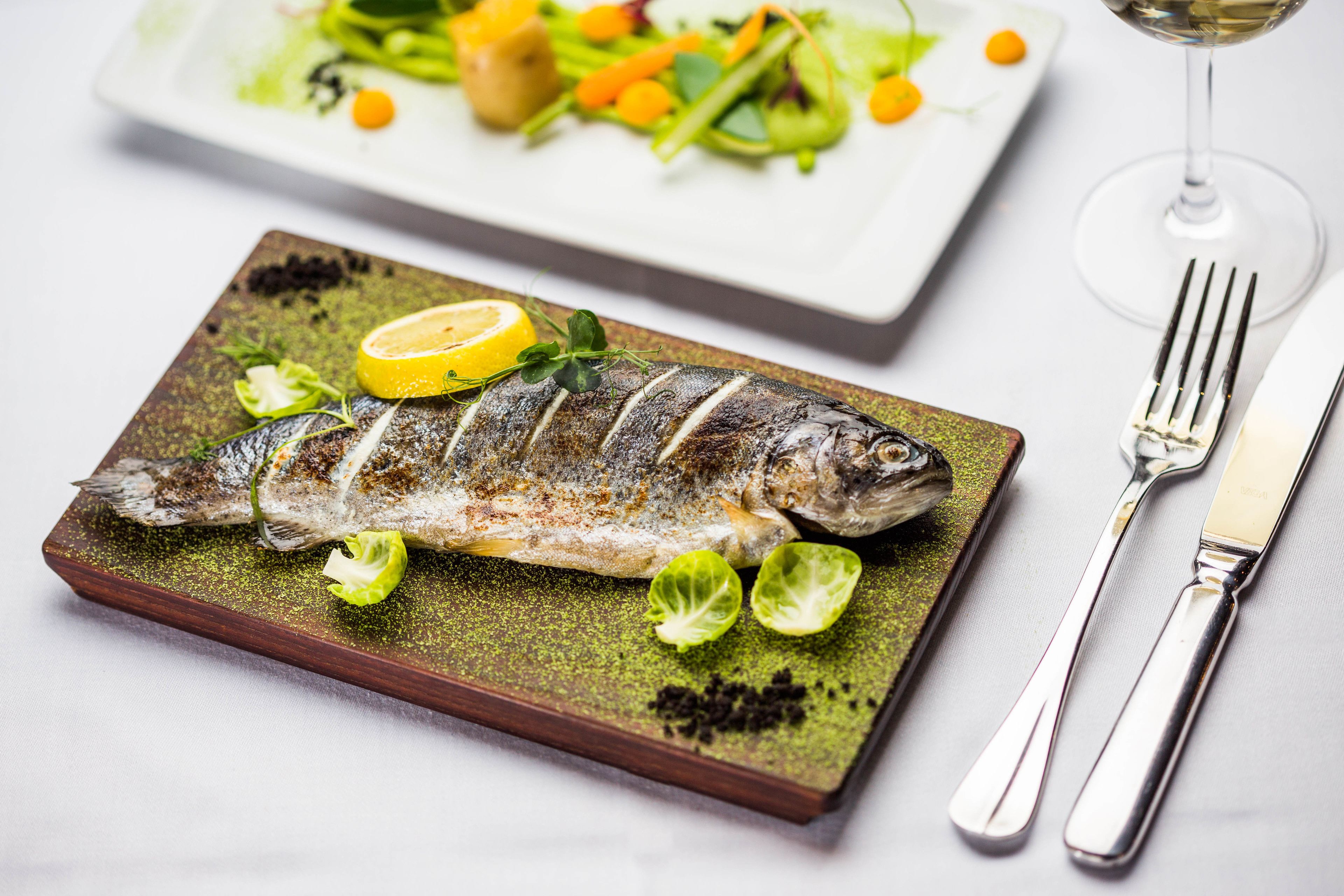A grilled fish on a plate with lemon and herbs, a fork and knife beside it, and a glass of white wine.