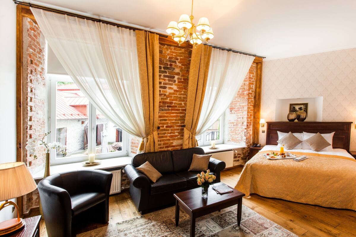 Une chambre d’hôtel avec un mur en briques, un canapé en cuir noir, une table basse en bois, un lit recouvert d’une courtepointe beige et un lustre.