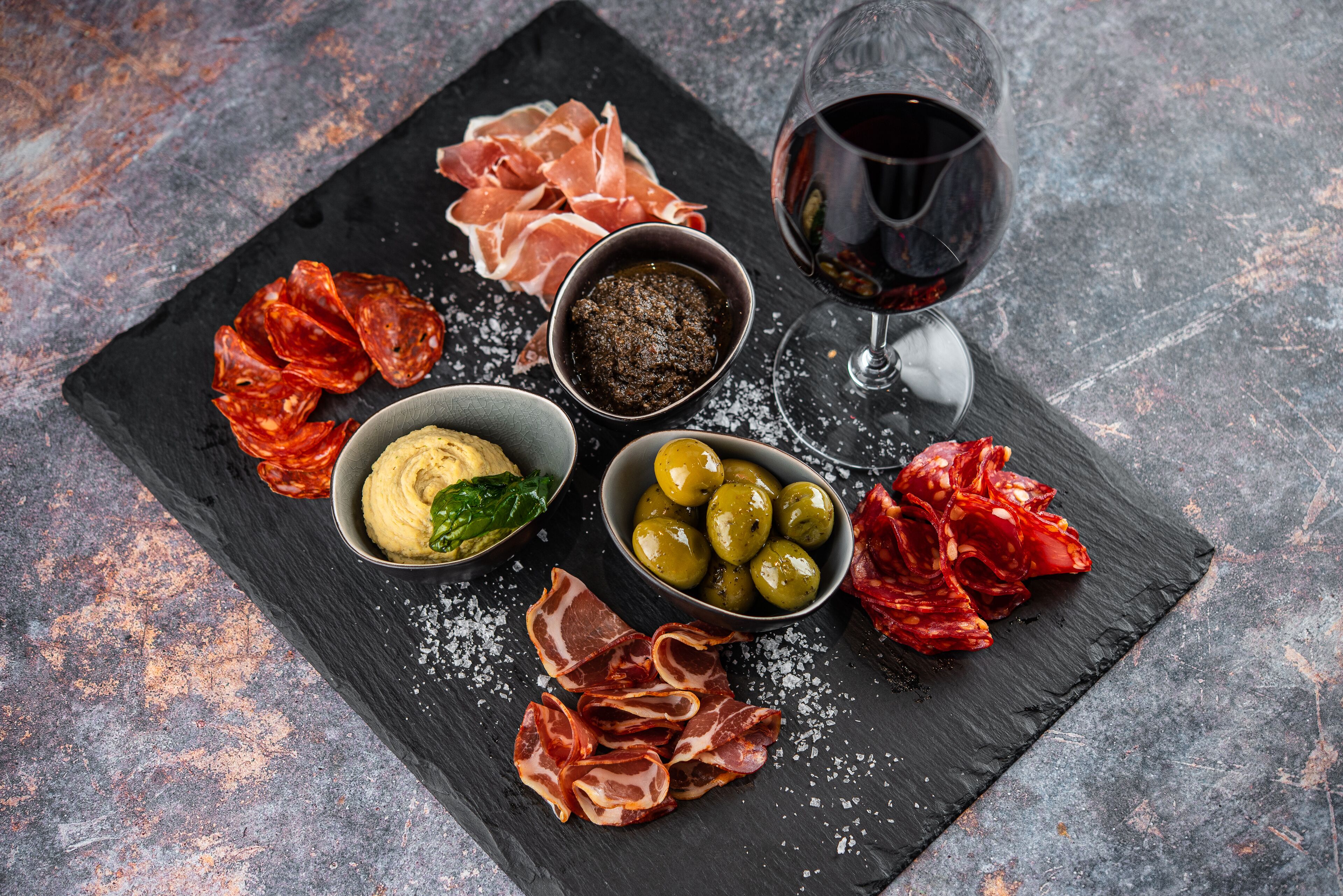 A black slate board with various food items including cured meats, olives, and a bowl of hummus, accompanied by a glass of red wine.