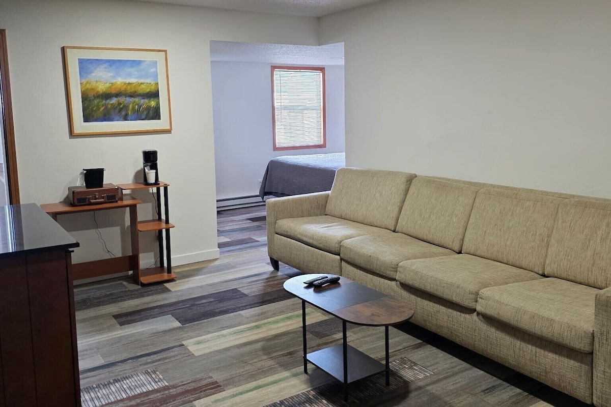 A living room with a beige sofa, a small coffee table, a desk with a computer, a painting on the wall, and a window with blinds.