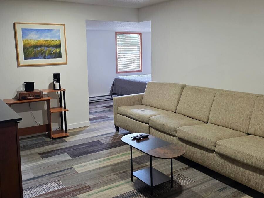 A living room with a beige sofa, a small coffee table, a desk with a computer, a painting on the wall, and a window with blinds.