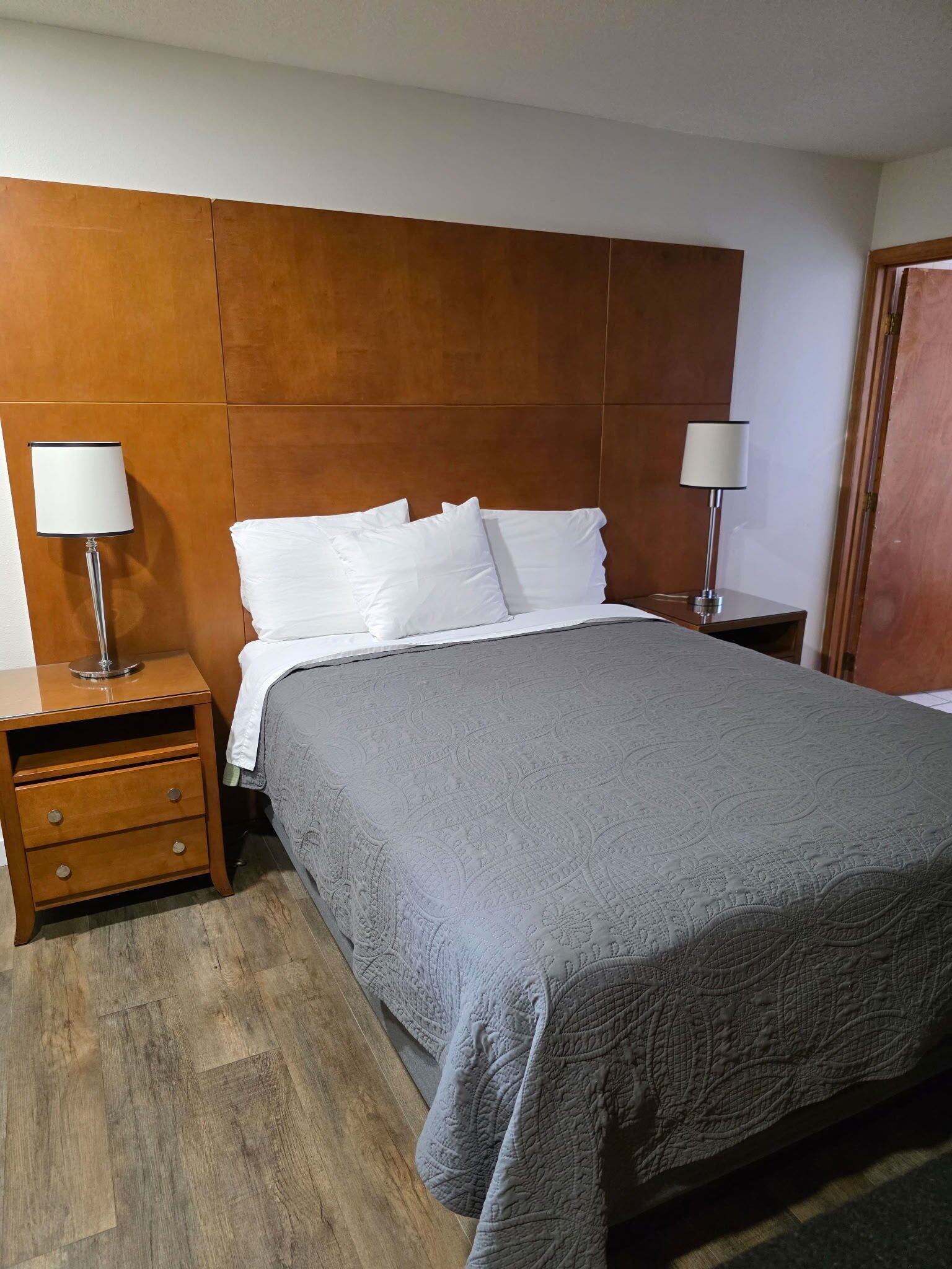 Uma cama bem arrumada com um edredom cinzento, duas mesas de cabeceira com abajures e uma cabeceira de madeira.