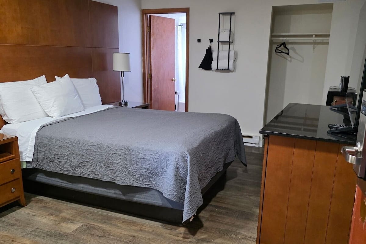 A bedroom with a bed, bedside table, lamp, and a small kitchenette area.