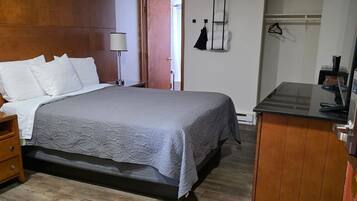 A bedroom with a bed, bedside table, lamp, and a small kitchenette area.