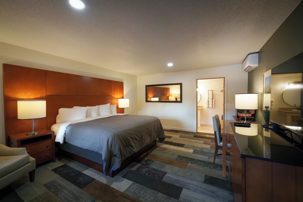 A hotel room with a large bed, bedside tables, a desk with a chair, a mirror, and a bathroom visible through an open door.
