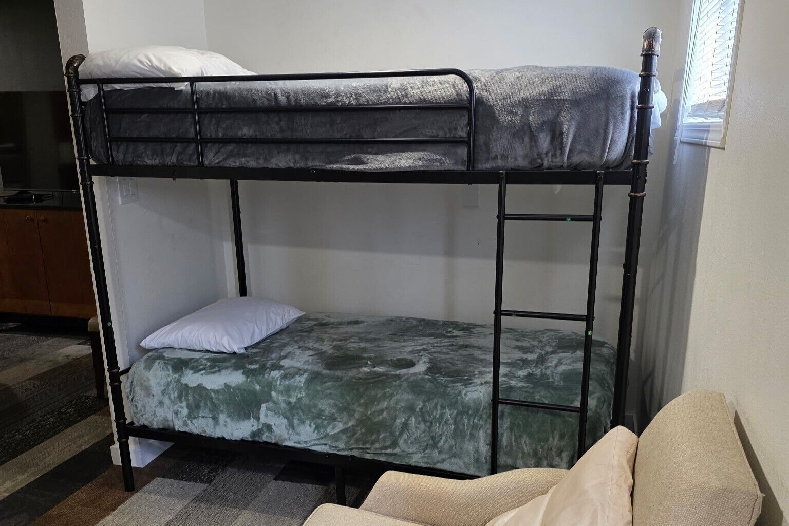 Una cama litera con colchón y almohadas, un aparador de madera y una silla en una habitación con ventana.