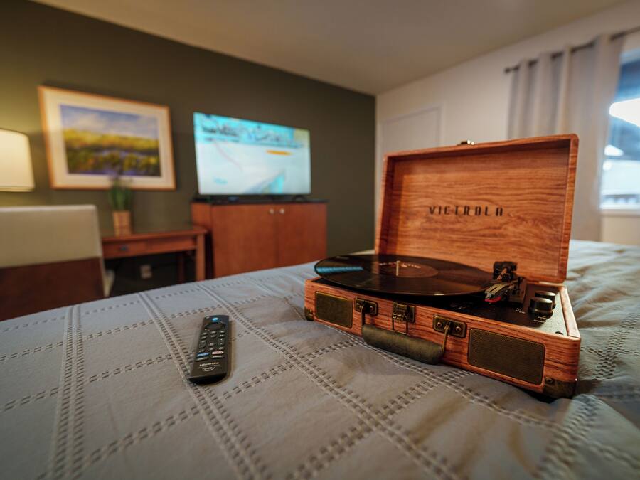 A wooden music player on a bed with a remote control beside it, a television on a stand, and a framed picture on the wall.