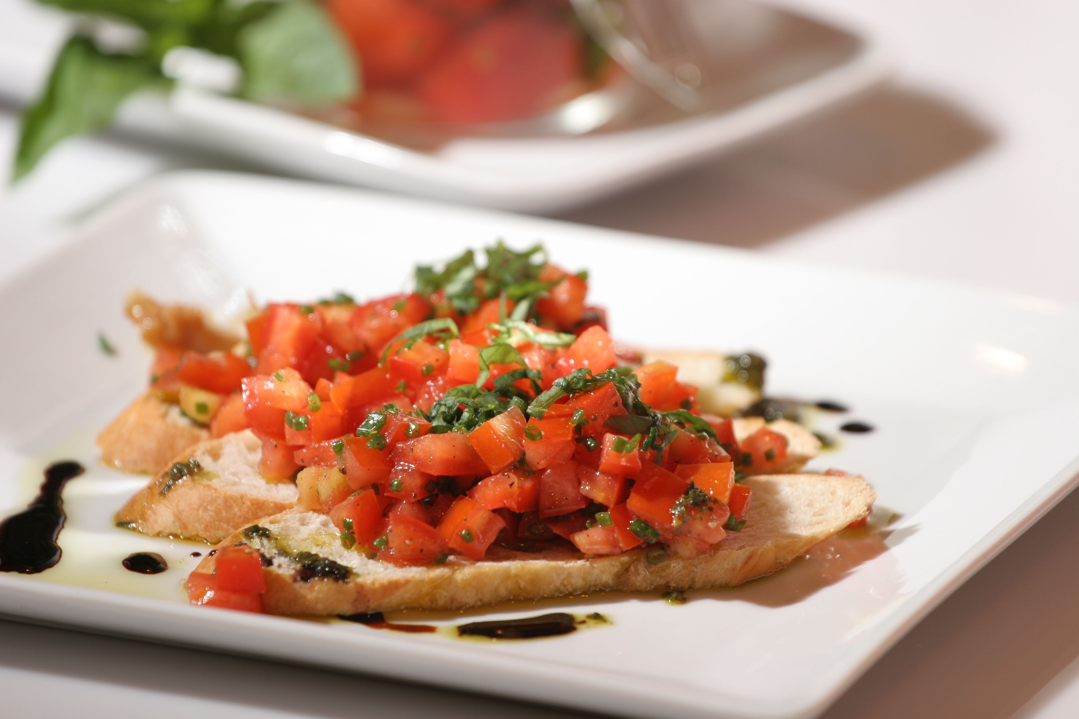 A square plate with a dish of bruschetta topped with diced tomatoes, herbs, and a drizzle of olive oil.