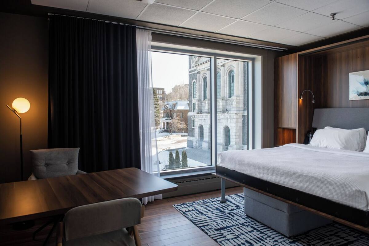 A hotel room with a large window, a bed, a desk, and a chair.
