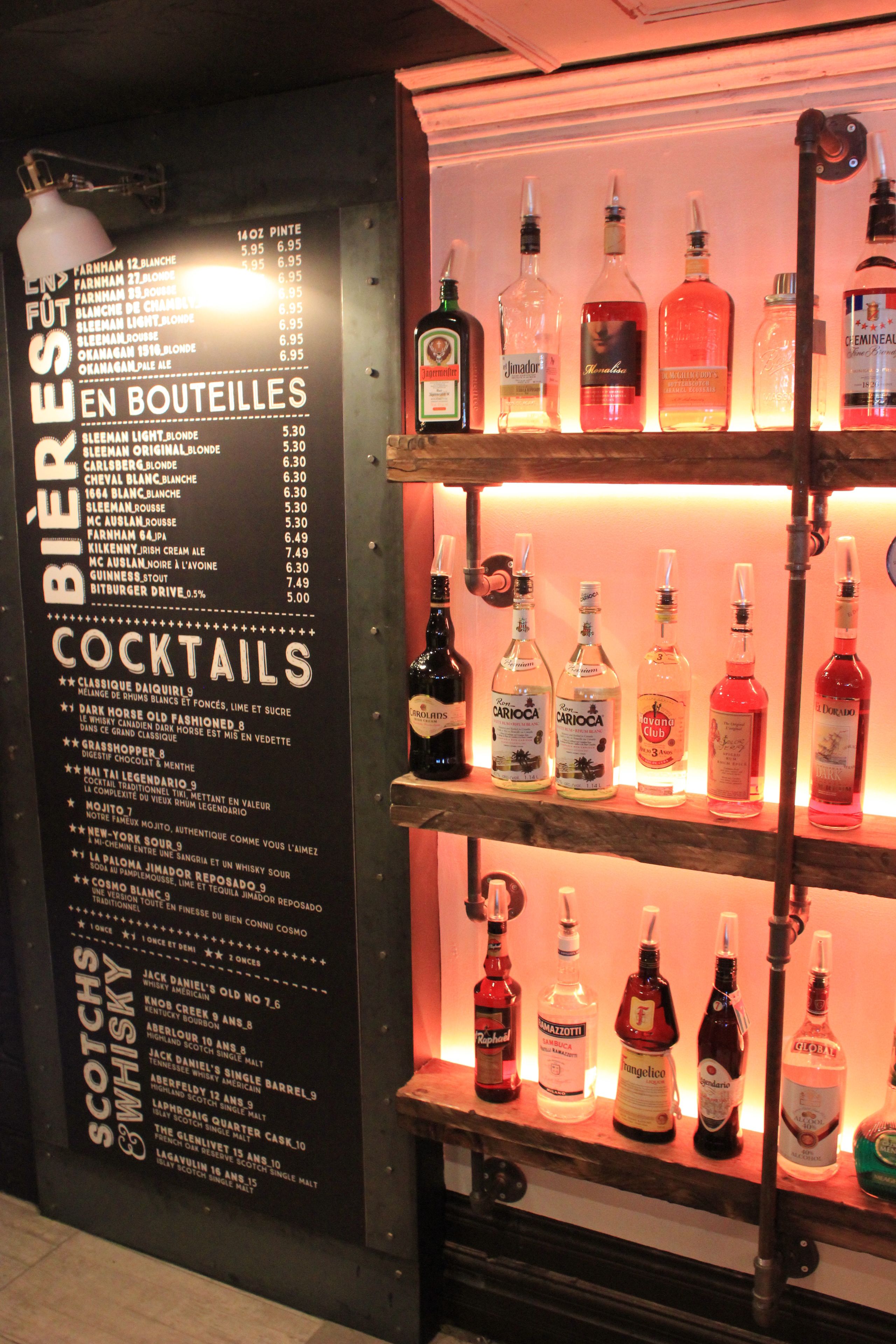 Uma área de bar com prateleiras cheias de garrafas de bebidas e um cardápio com diversos drinques.