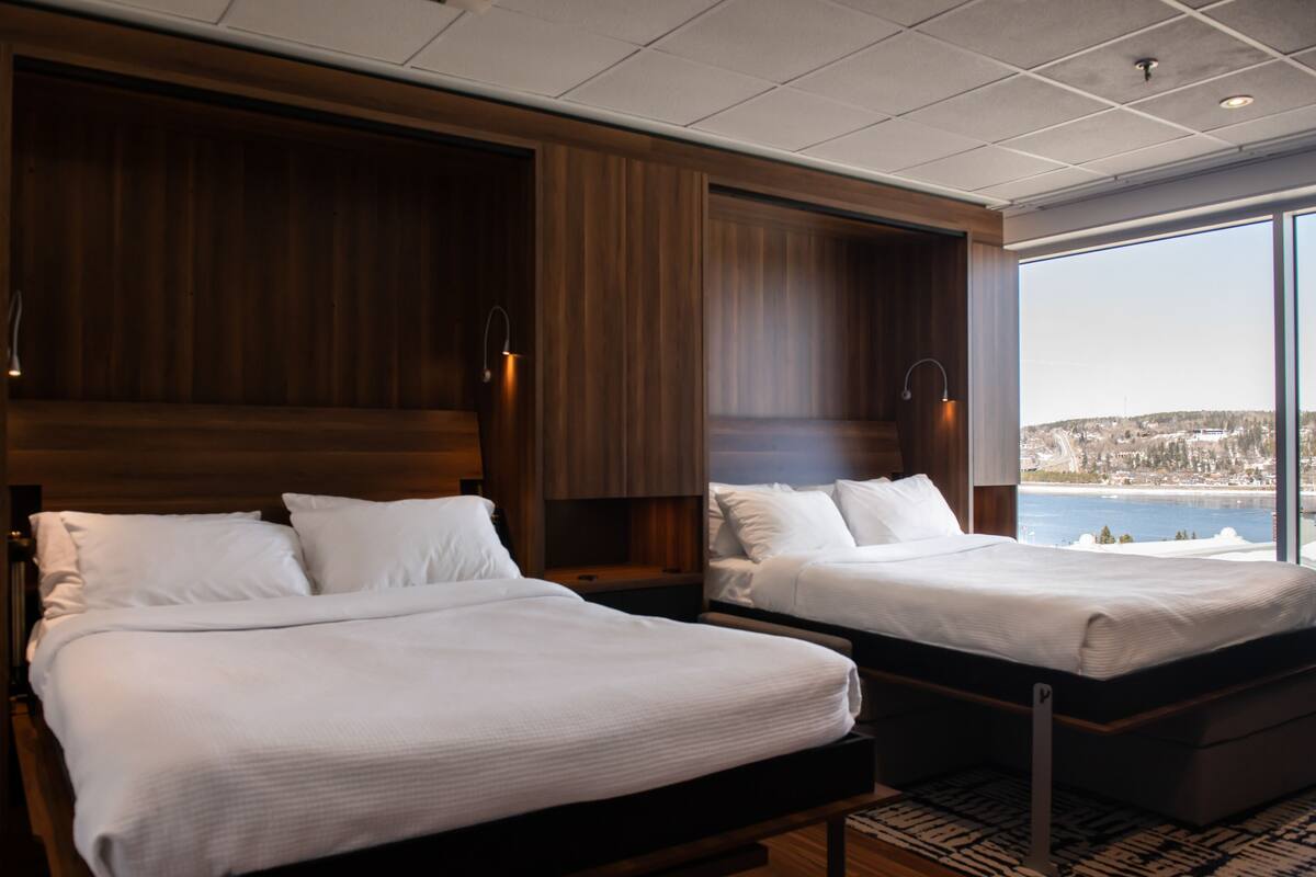 A hotel room with two beds, wooden headboards, and a large window offering a view of a body of water and a cityscape.