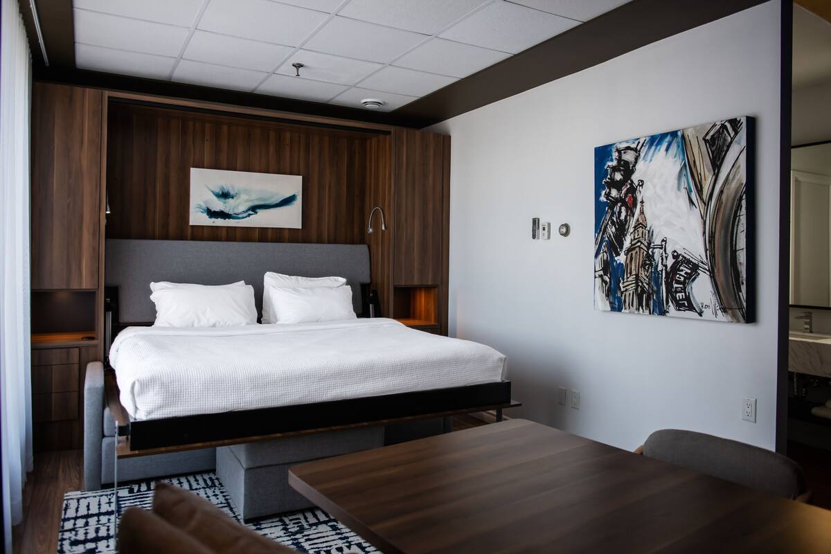 A modern hotel room with a large bed, a wooden headboard, a painting on the wall, and a dining table.