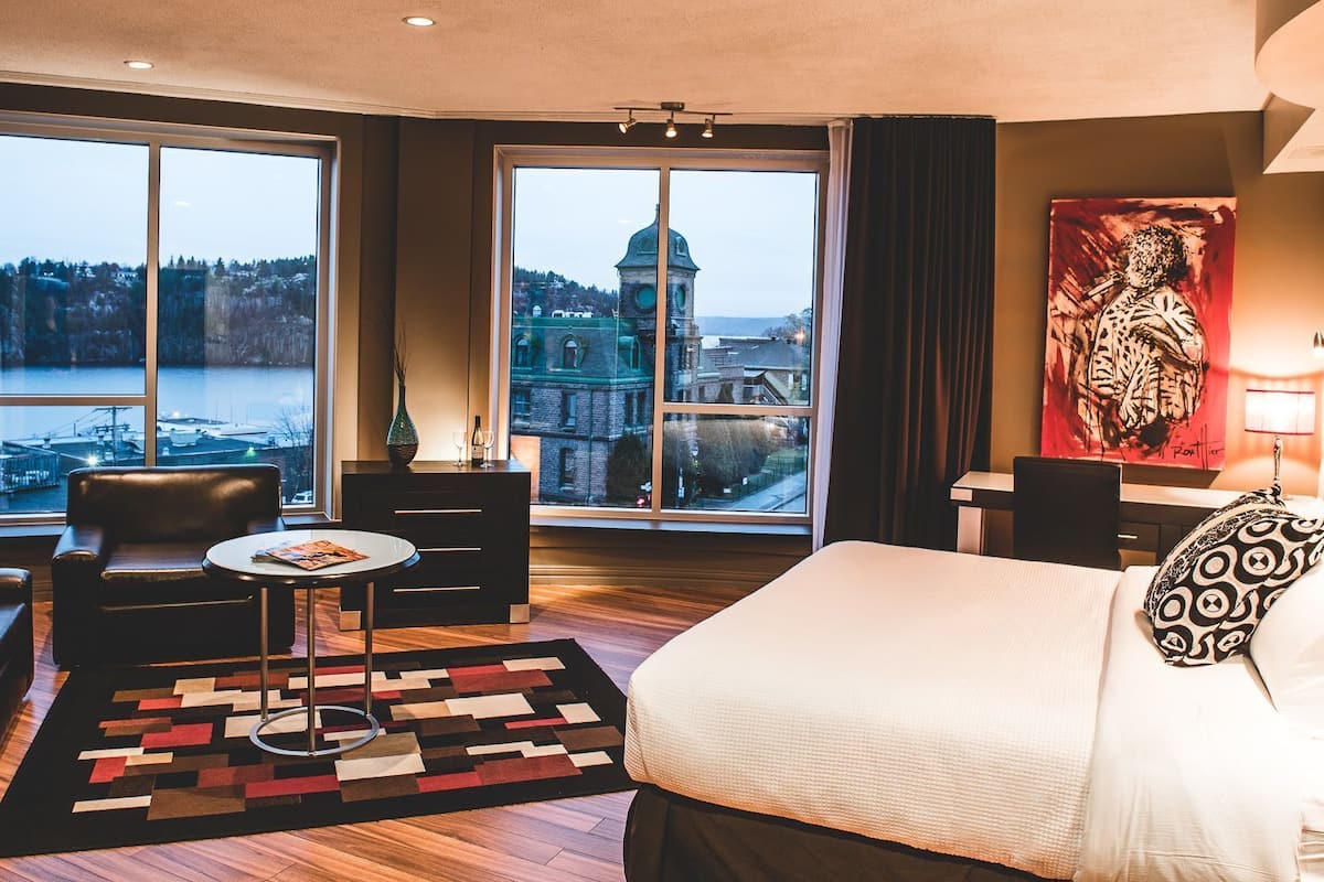 A hotel room with a large bed, a desk, a chair, a small table, and two windows offering a view of a cityscape and a body of water.
