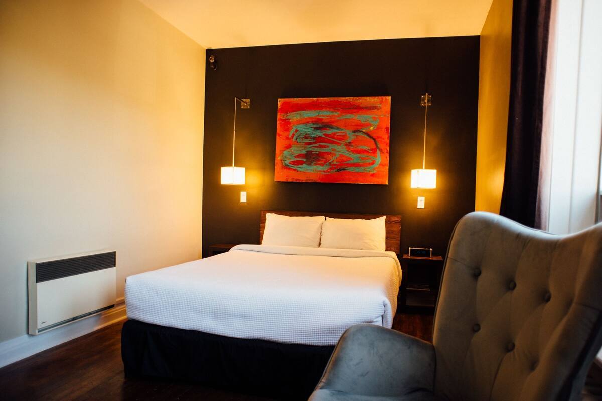 A hotel room with a bed, a chair, a wall-mounted artwork, and two wall sconces.