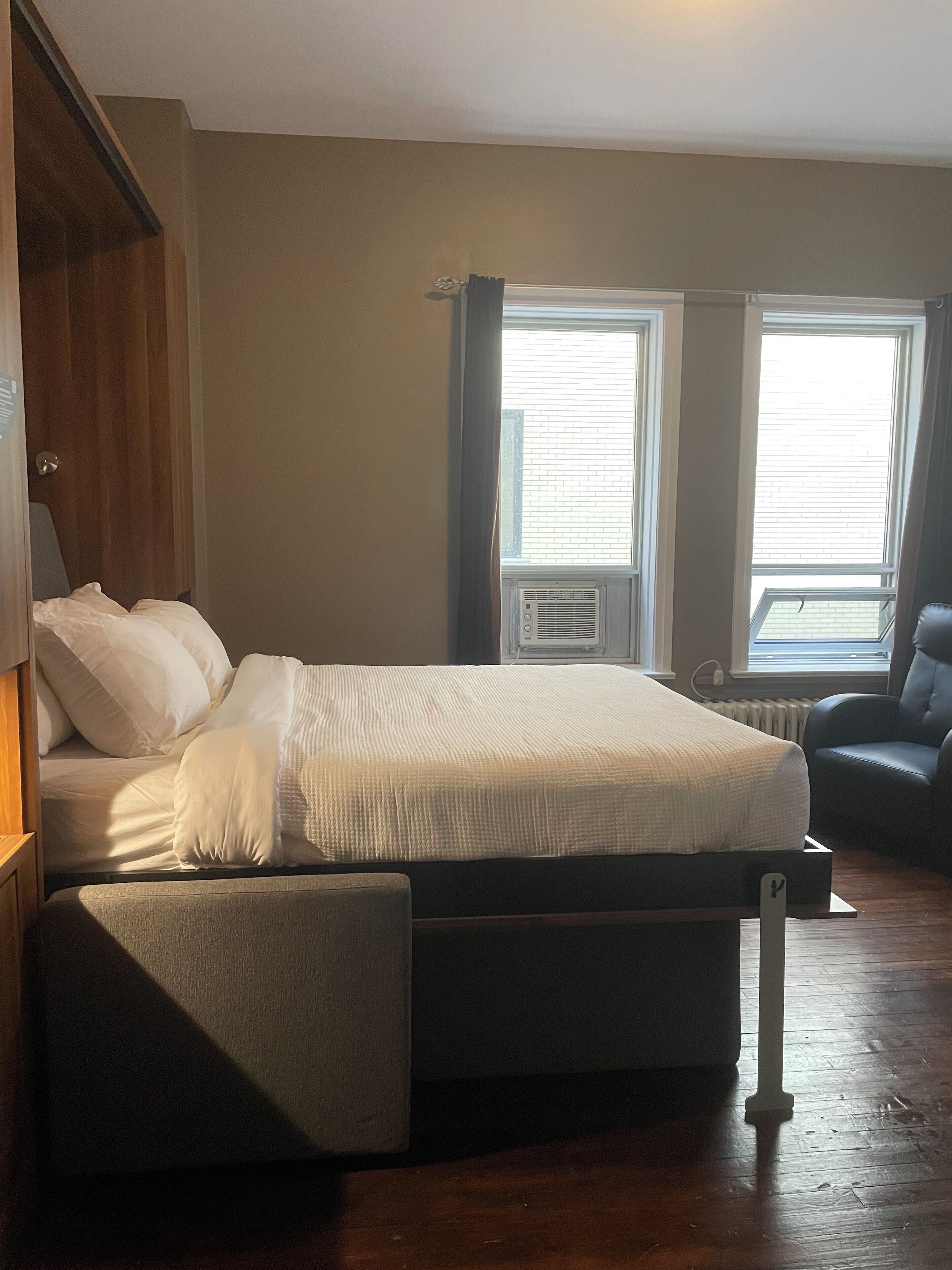 Un dormitorio con cama, una silla y dos ventanas con cortinas.