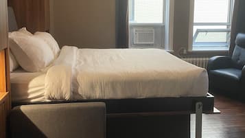 Un dormitorio con cama, una silla y dos ventanas con cortinas.