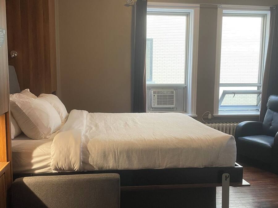 Un dormitorio con cama, una silla y dos ventanas con cortinas.