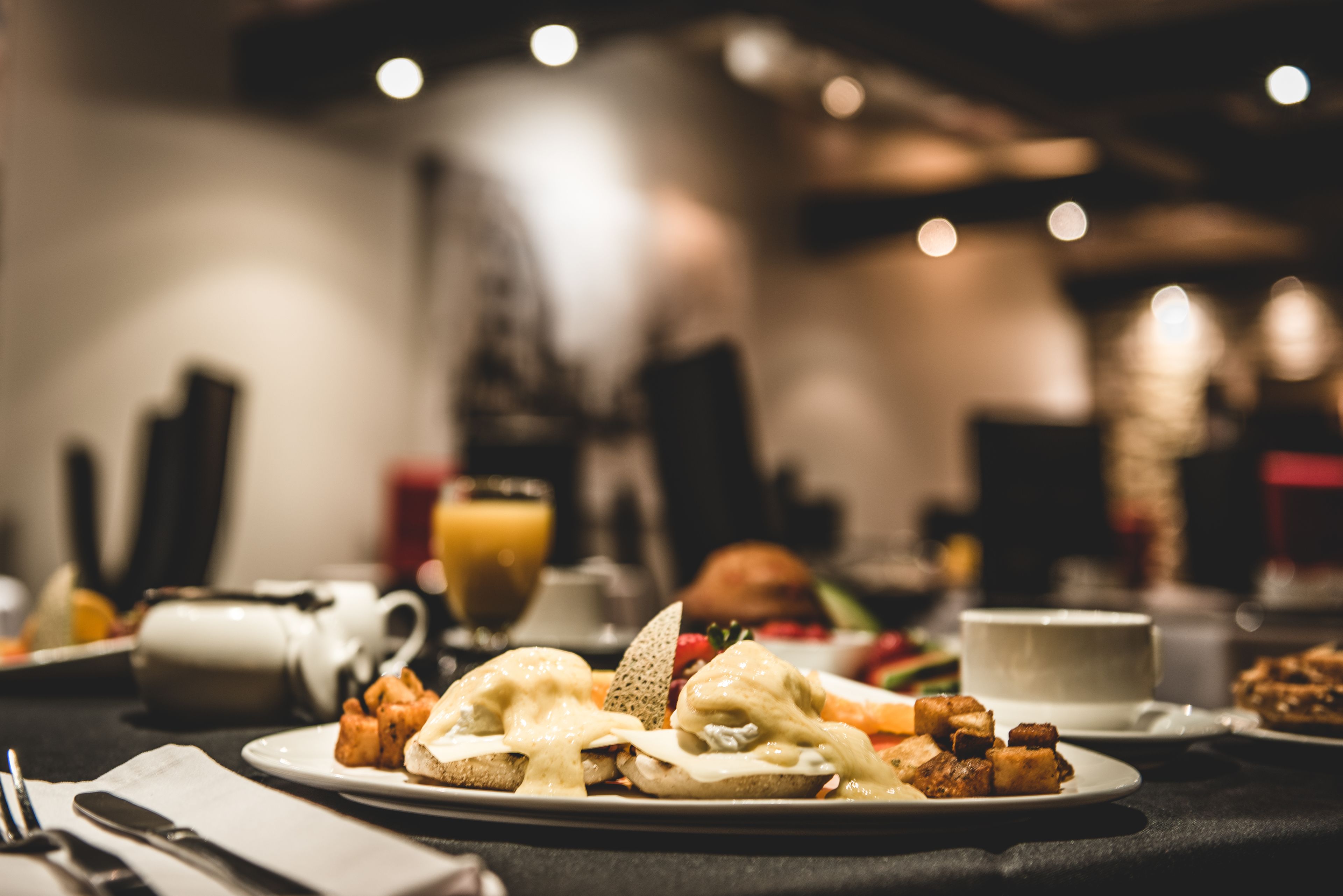 Une table mise pour le petit-déjeuner avec une assiette d’œufs Bénédicte, des toasts, des fruits, une tasse de café et un verre de jus d’orange.