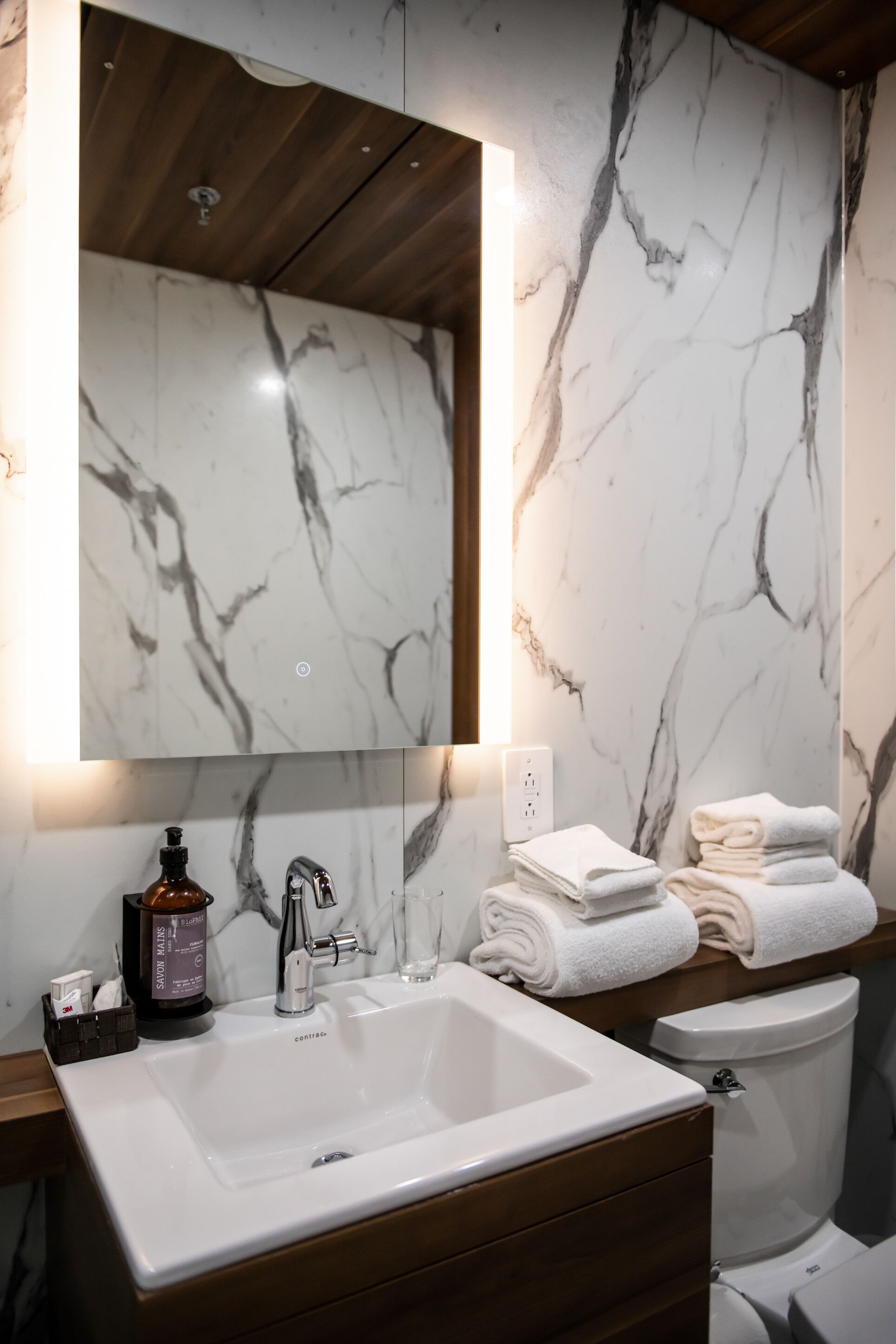 Un baño con pared de mármol, un lavamanos blanco, un espejo y toallas apiladas.