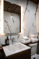 Un baño con pared de mármol, un lavamanos blanco, un espejo y toallas apiladas.