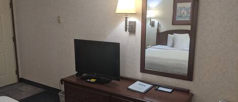 A hotel room with a bed, a TV on a wooden dresser, a lamp, a mirror, and a small desk with a phone and a certificate.
