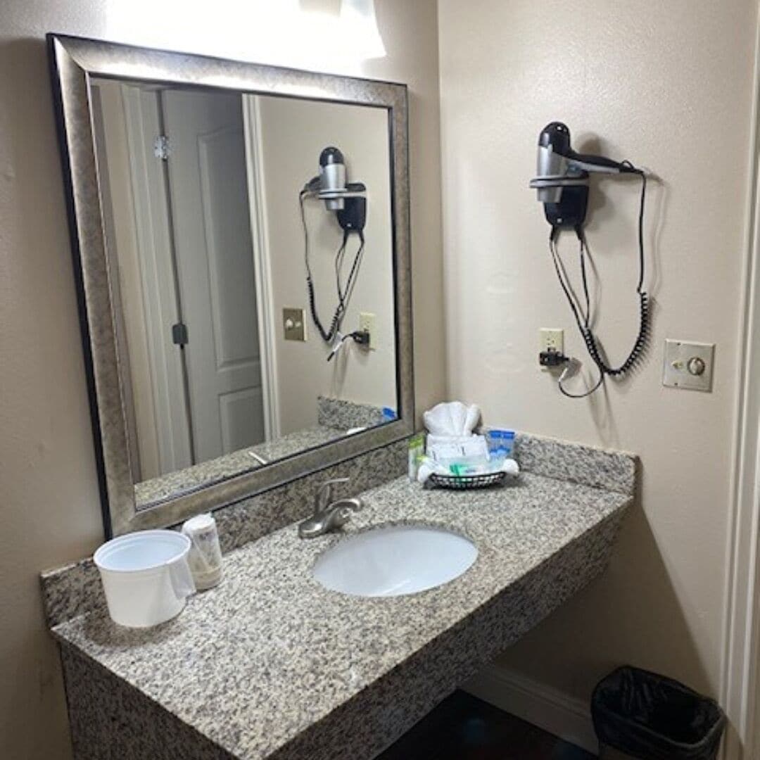 A bathroom with a granite countertop, a sink, a mirror, a hairdryer, and a trash can.