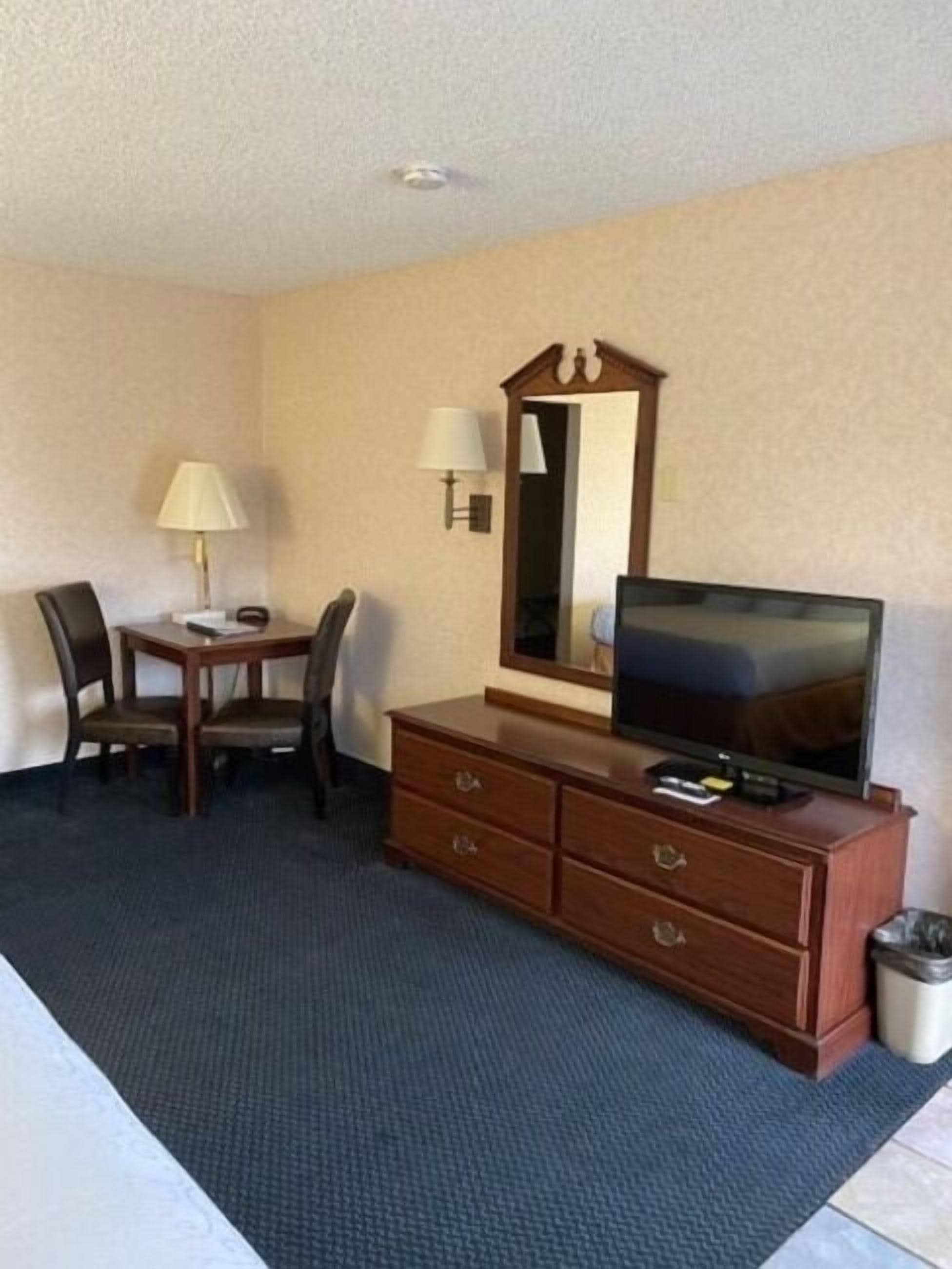 A hotel room with a wooden dresser, a television, a desk with two chairs, a lamp, and a mirror.