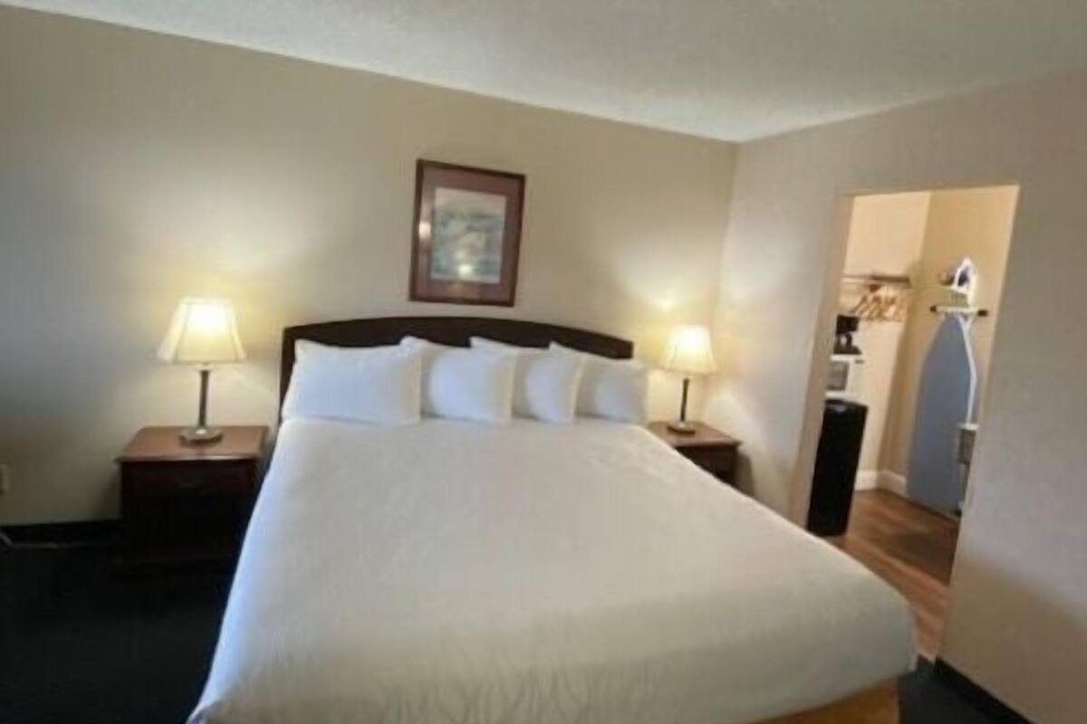 A neatly made bed with white linens and a brown skirt, two bedside tables with lamps, a framed picture on the wall, and a doorway leading to another room.