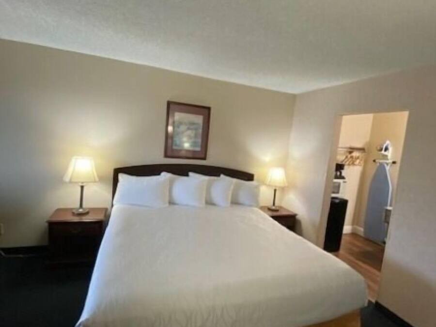 A neatly made bed with white linens and a brown skirt, two bedside tables with lamps, a framed picture on the wall, and a doorway leading to another room.