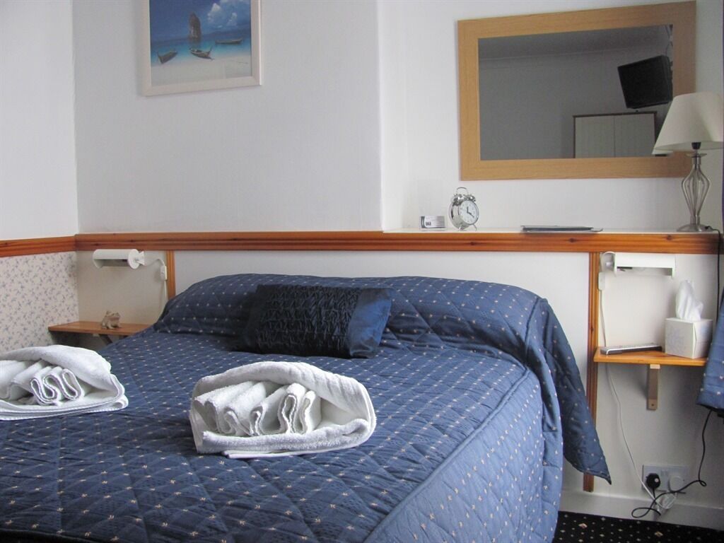 A neatly made bed with a blue quilt, two rolled towels, a bedside table with a lamp, a small clock, and a framed picture on the wall.
