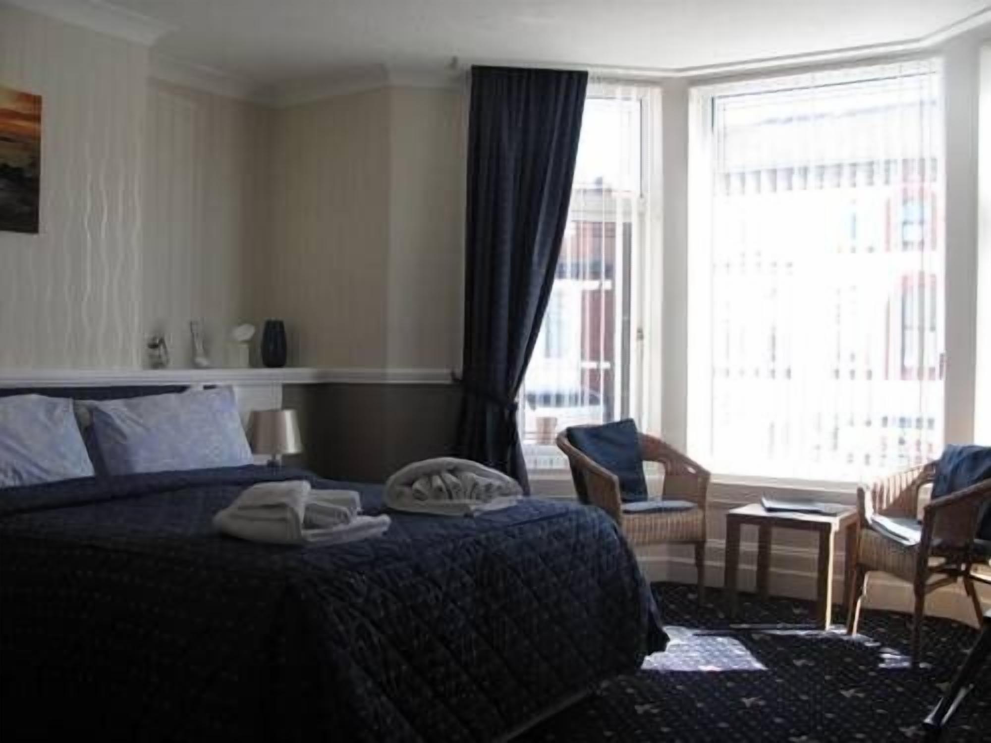 A hotel room with a large bed, a small table, two chairs, a window with curtains, and a view of buildings outside.