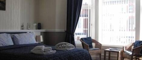 A hotel room with a large bed, a small table, two chairs, a window with curtains, and a view of buildings outside.