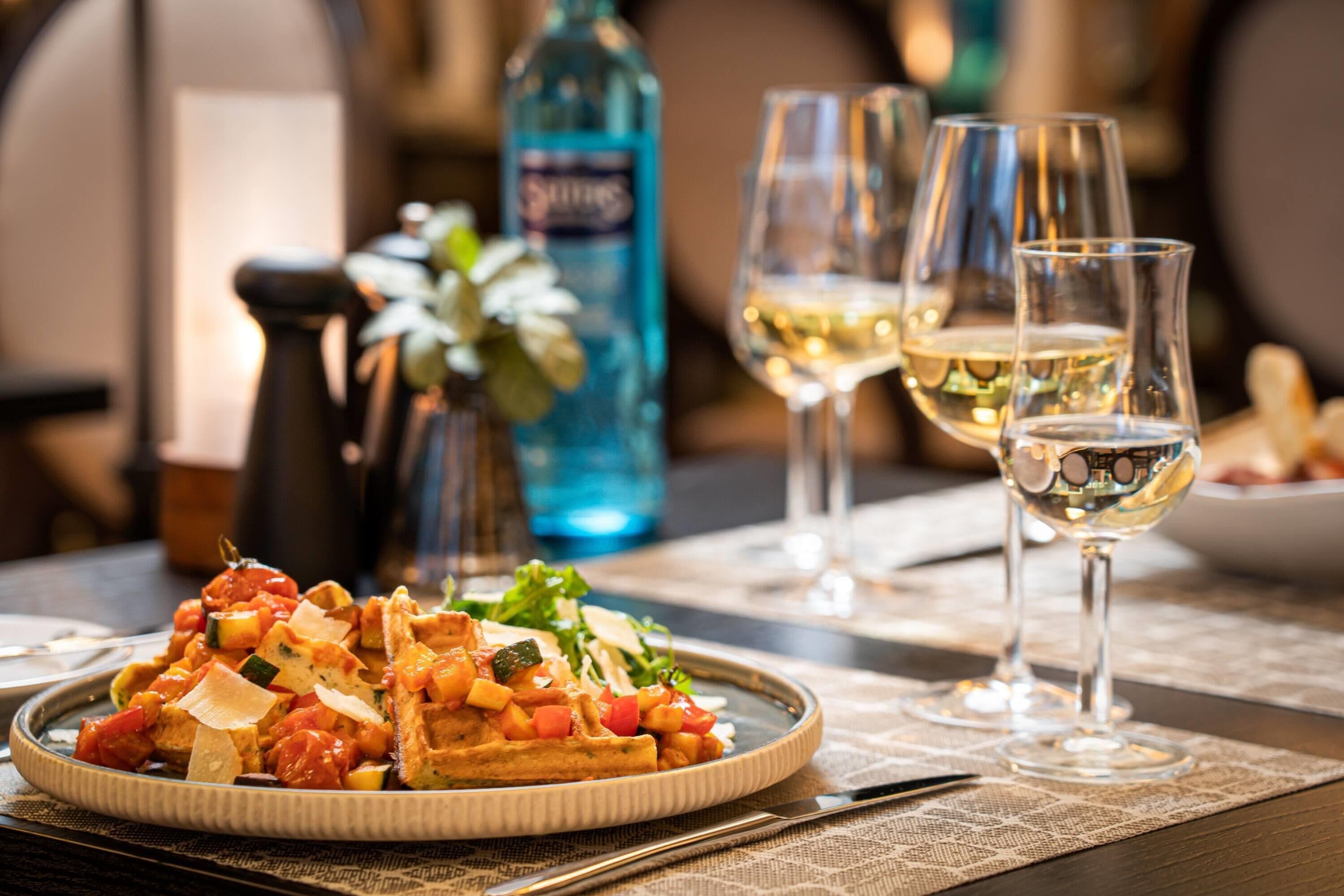 Une table à manger avec une assiette de nourriture, des verres de vin blanc et une bouteille d’eau.