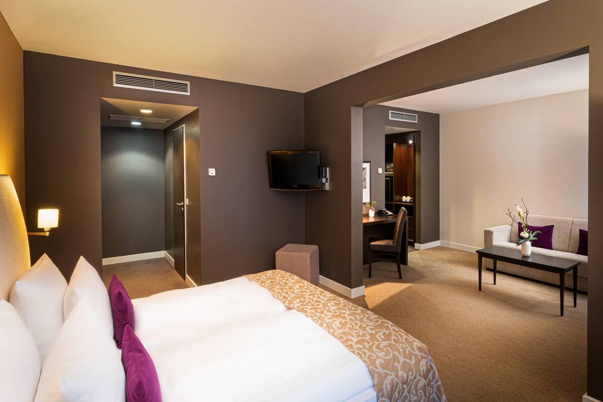 A hotel room with a bed, a desk, a television, and a seating area.