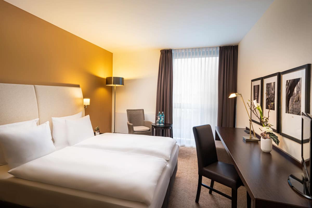 A hotel room with a large bed, a desk, a chair, a lamp, and framed pictures on the wall.
